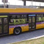 A imagem mostra um ônibus elétrico amarelo, com listras roxas e adesivos com a frase "100% elétrico – energia limpa carioca".