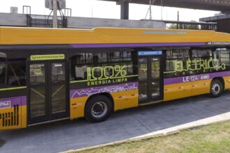A imagem mostra um ônibus elétrico amarelo, com listras roxas e adesivos com a frase "100% elétrico – energia limpa carioca".