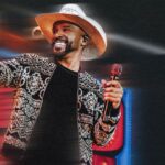 Cantor Alexandre Pires com chapéu de palha e jaqueta estampada sorrindo enquanto segura microfone