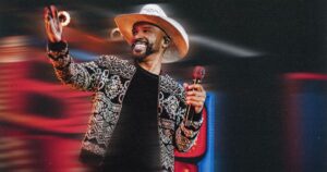 Cantor Alexandre Pires com chapéu de palha e jaqueta estampada sorrindo enquanto segura microfone