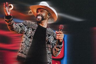 Cantor Alexandre Pires com chapéu de palha e jaqueta estampada sorrindo enquanto segura microfone