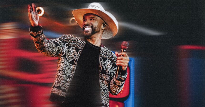 Cantor Alexandre Pires com chapéu de palha e jaqueta estampada sorrindo enquanto segura microfone