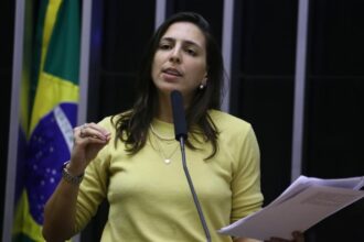 Natália Bonavides veste camisa de manga amarela, colares e relógio. É branca e tem cabelo médio castanho escuro.