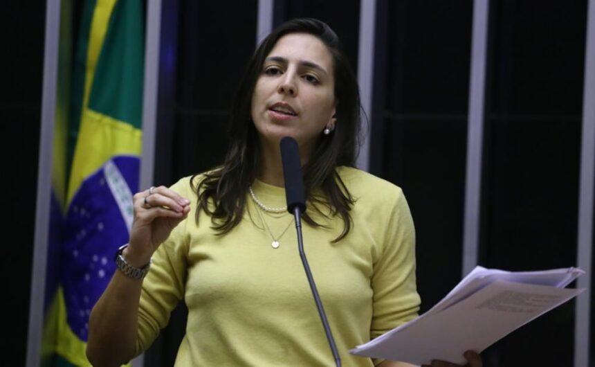 Natália Bonavides veste camisa de manga amarela, colares e relógio. É branca e tem cabelo médio castanho escuro.